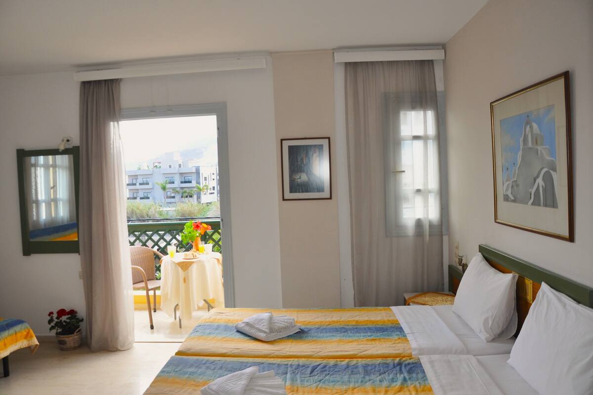 A hotel room with two beds, a balcony with a table and chairs, and a view of buildings outside.