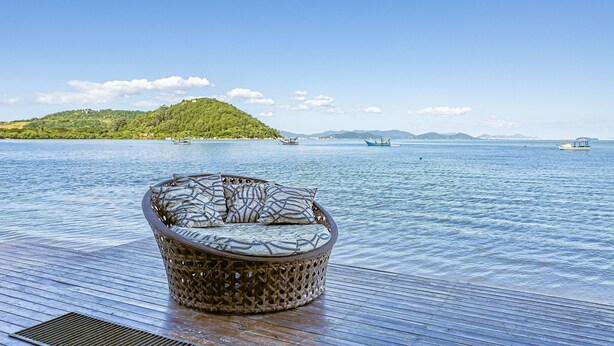 Un fauteuil de relaxation en osier, placé sur une terrasse en bois donnant sur une étendue d’eau calme, avec des bateaux et une île au loin.