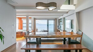 Un espace repas moderne avec une grande table et des chaises en bois, un luminaire suspendu contemporain et une vue sur la mer grâce à de grandes fenêtres.