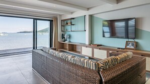 Un salon moderne doté d’une grande fenêtre donnant sur un plan d’eau, d’un téléviseur à écran plat fixé au mur et d’un meuble de rangement intégré pour les appareils multimédias.