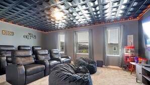 A home theater room with a ceiling grid, reclining chairs, bean bags, and a red cart with snacks.