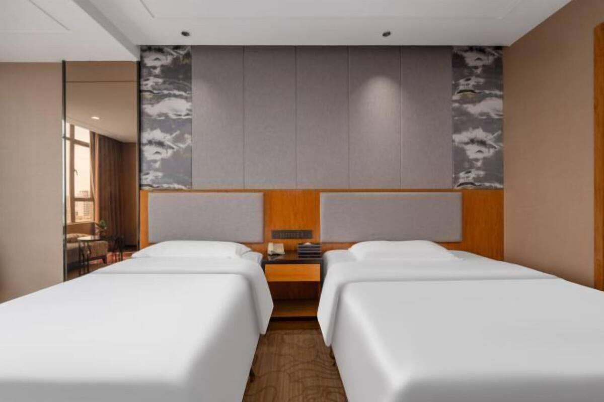 Habitación de hotel con dos camas, cabecera de madera, una mesita con lámpara y vistas al exterior.