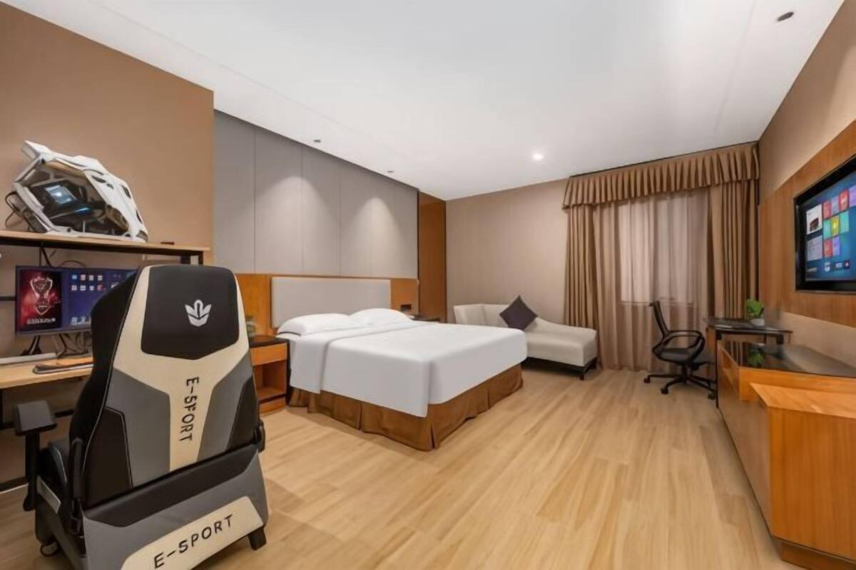 Habitación de hotel con cama, escritorio con computadora, silla gamer, televisor y mostrador de recepción.