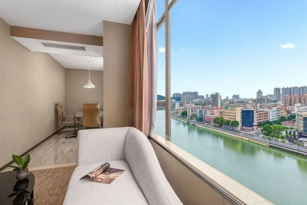 Habitación de hotel moderna con un ventanal grande, un sofá, una mesa con una planta y vistas a un paisaje urbano y un río.