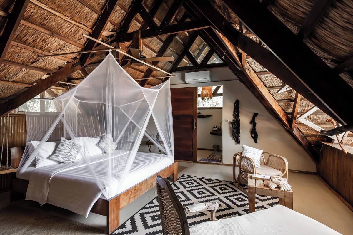 A bedroom with a bed under a thatched roof, a wooden bench, and a patterned rug.