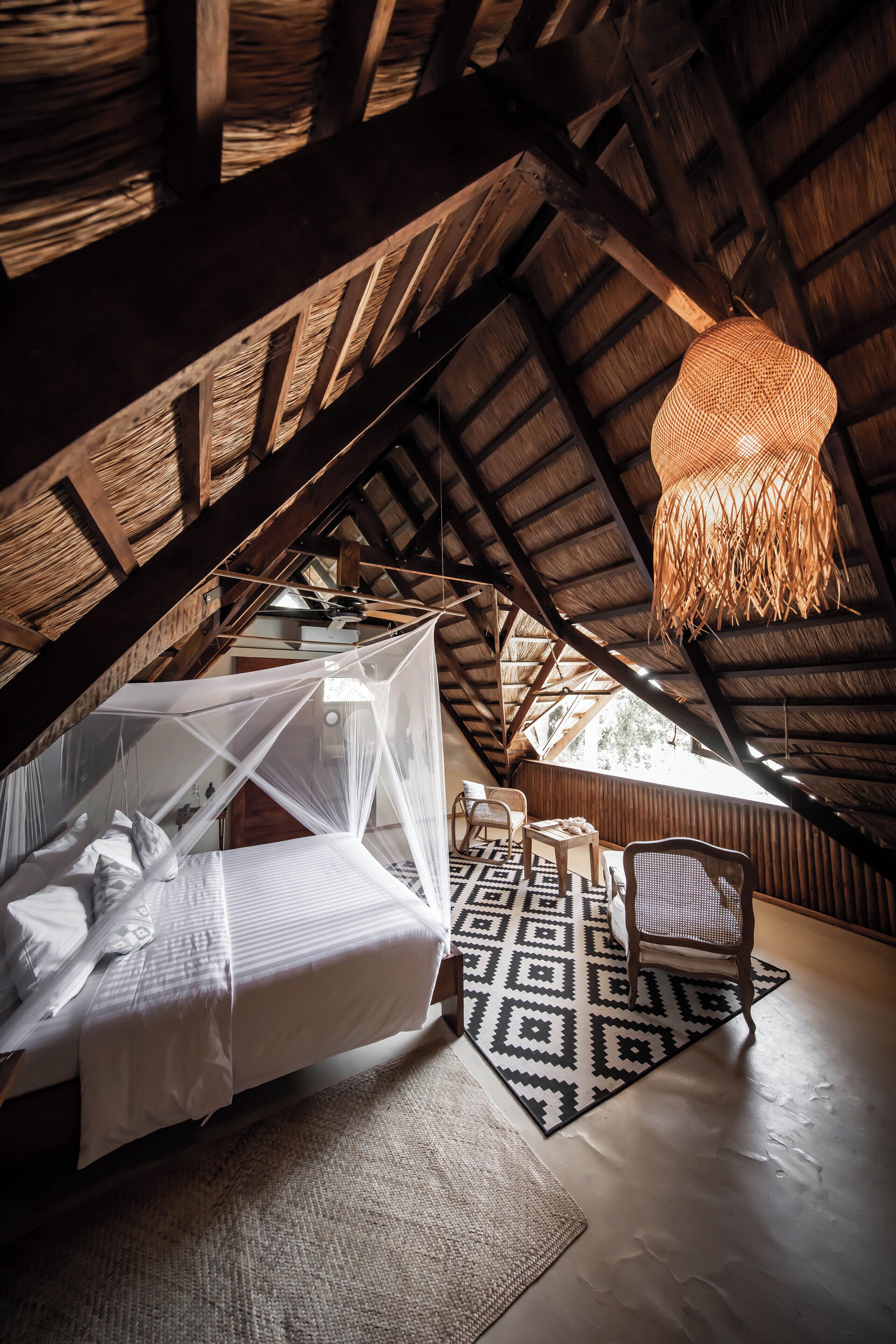 A high-ceilinged attic bedroom with a bed, a woven hanging light fixture, and a patterned rug.