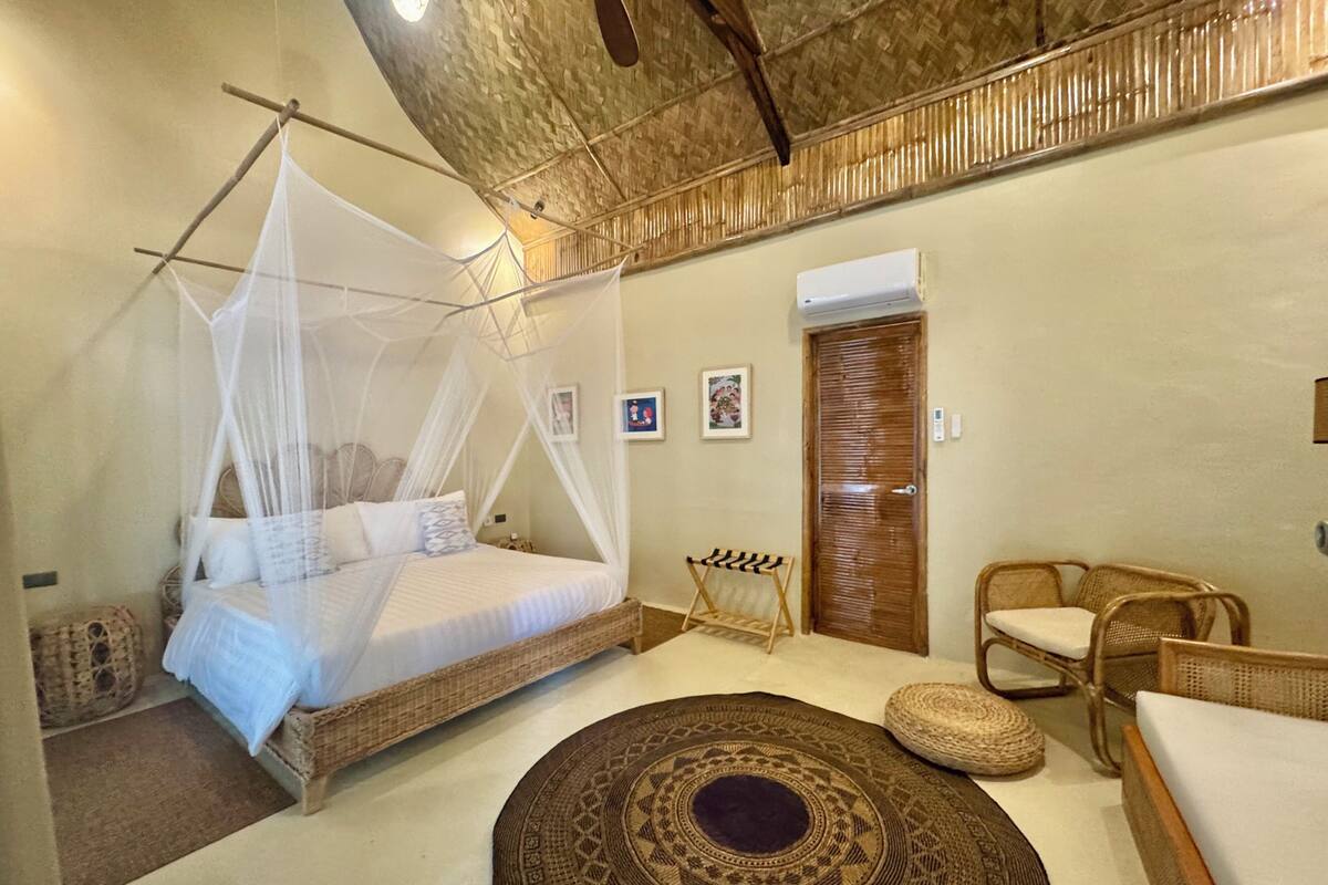 A room with a bed under a mosquito net, a wooden door, wicker furniture, and a patterned rug.