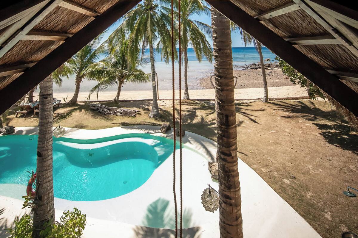 A beach view with a pool, palm trees, and a thatched roof structure.
