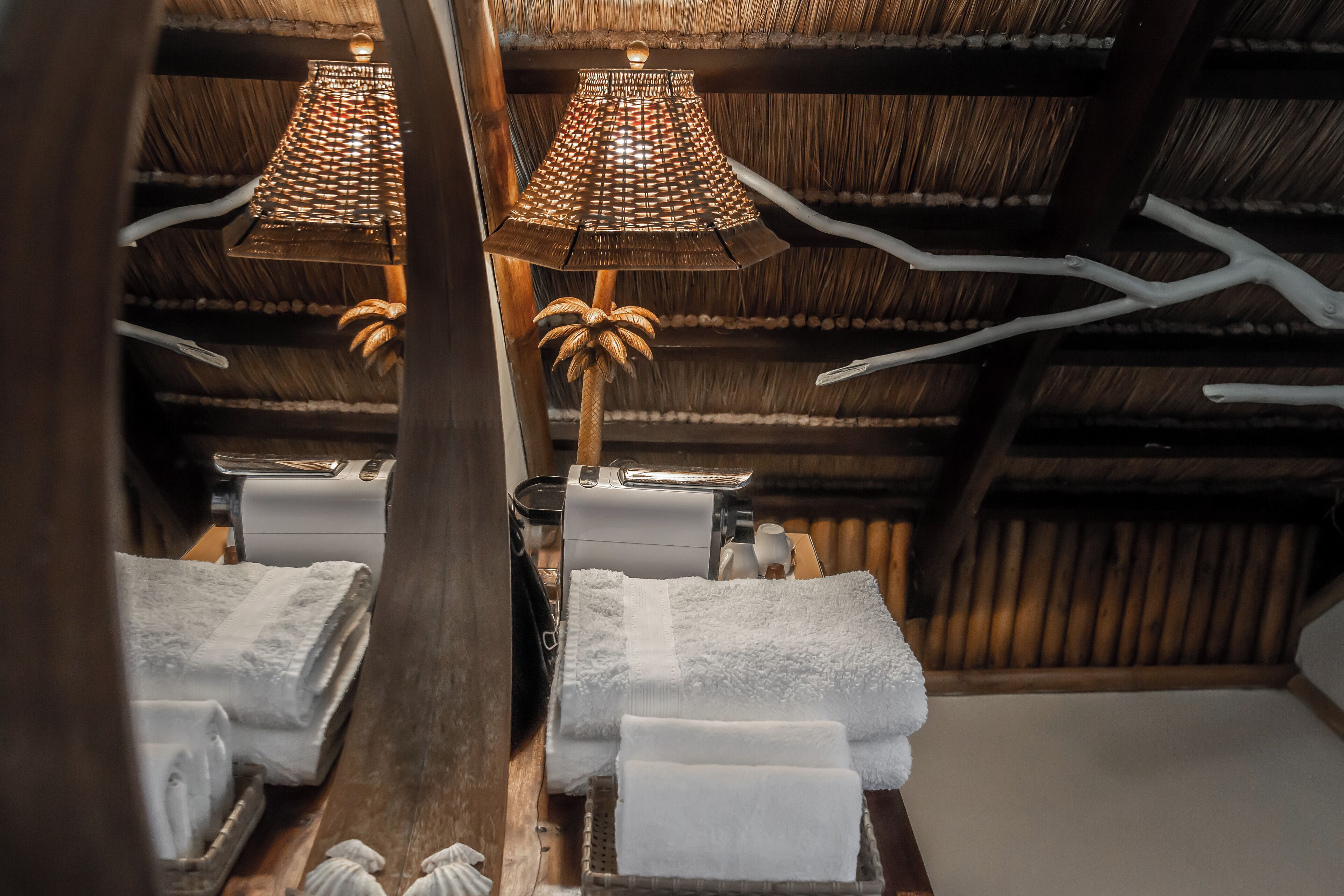 Une pièce avec un plafond en chaume, un plancher en bois et un lit recouvert de serviettes blanches. On y trouve deux lampes dotées d’un abat-jour en osier et d’un pied en bois.