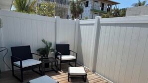 Eine Terrasse im Freien mit Holzboden, zwei dunkle Korbstühle, ein kleiner Tisch mit einer Pflanze und eine weiße gepolsterte Bank.