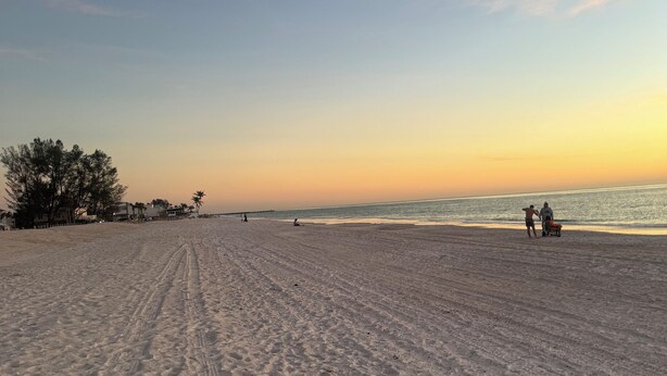 Ein weiter Sandstrand mit Fußabdrücken, einigen Personen und einem Sonnenuntergang im Hintergrund.