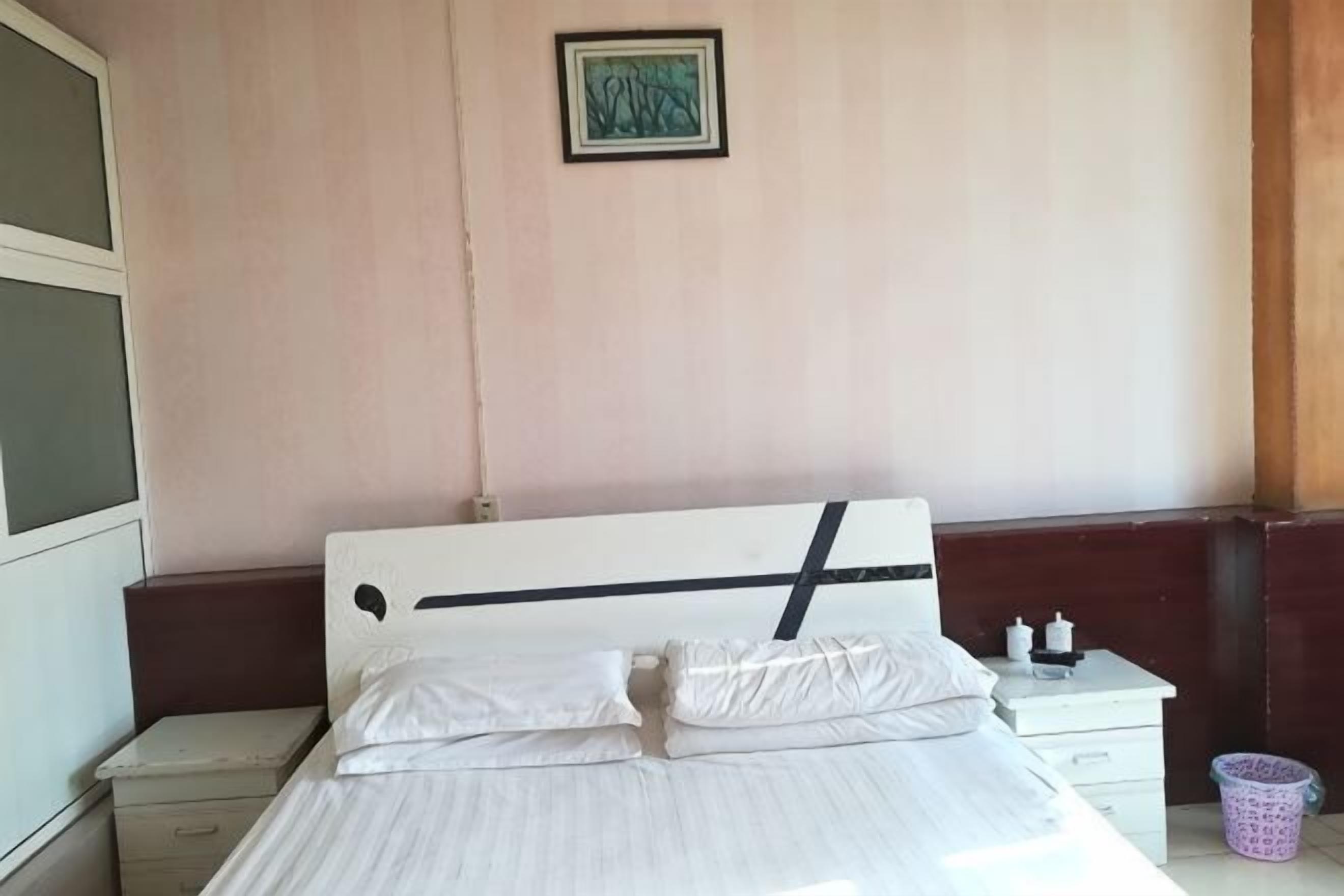 Una cama con ropa de cama blanca, una mesita de noche con lámpara y un cubo de basura pequeño.