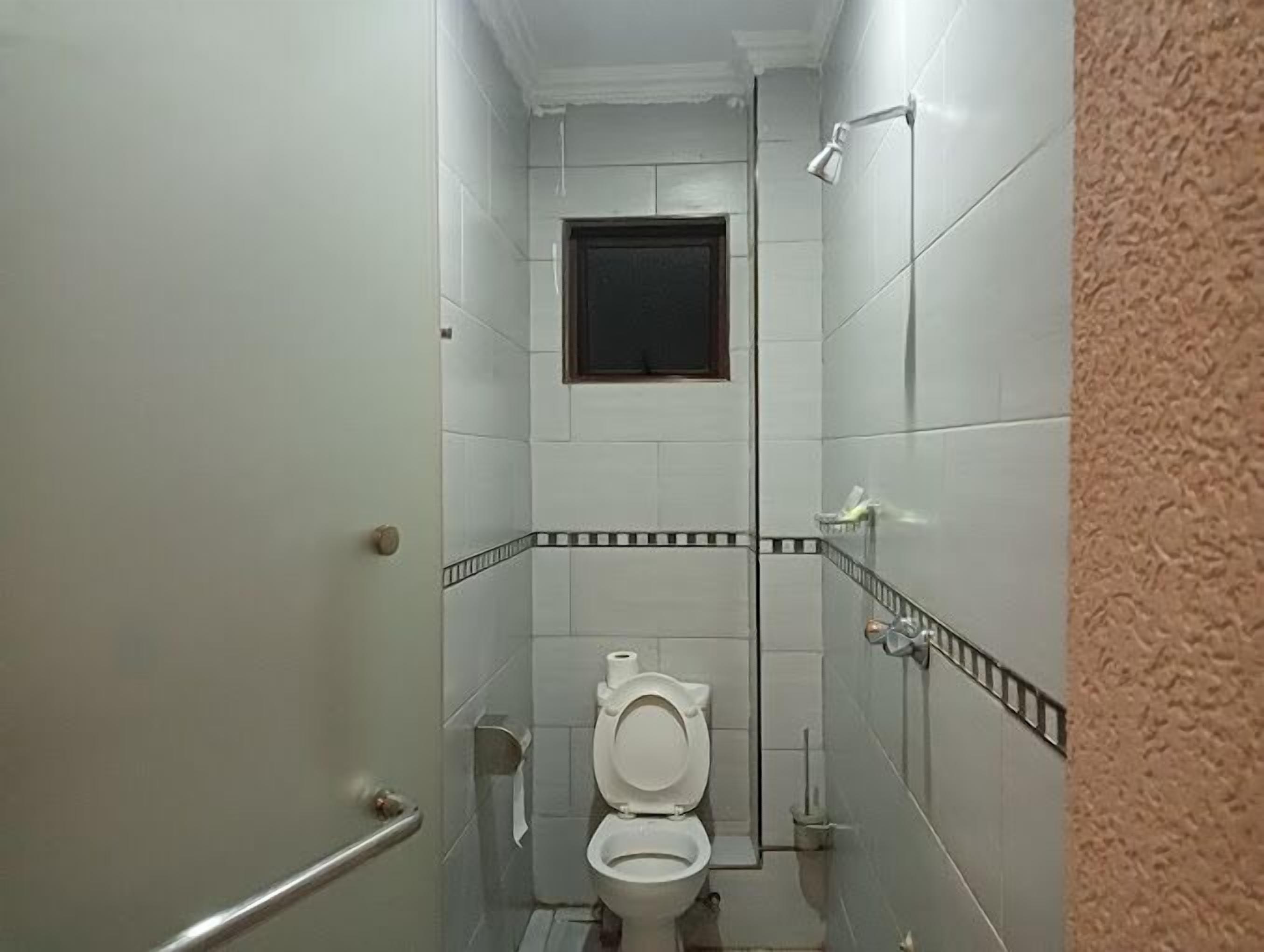 A small, narrow bathroom with white tiles, a toilet, a shower, and a small window.