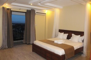 A hotel room with a large bed, a wooden headboard, and a view through a glass door. - Rockwell Hotel (Kisumu)