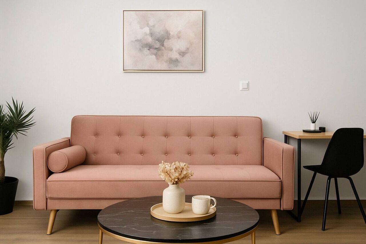 A pink sofa with a vase and mug on a round table, a small desk with a chair, and a plant on the left.