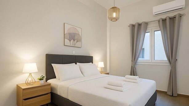 A neatly made bed with white linens, a bedside table with a lamp, and a wall-mounted air conditioner.