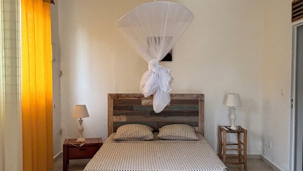 Un dormitorio con cama, ventilador de techo, mesita de noche con lámpara y ventana con cortinas.