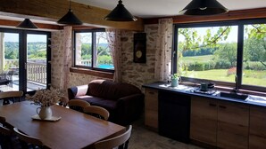 A cozy kitchen with a large window, a dining area with a table and chairs, and a stone wall.