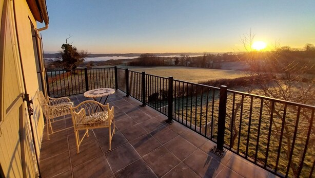 A balcony with a view of a lake and sunset. There are two chairs and a small table.