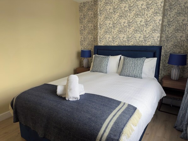 Een netjes opgemaakt bed met een blauw hoofdeinde, witte lakens en een blauw-wit gestreept deken. Er zijn twee nachtkastjes met lampen en een muur met bloemenbehang.