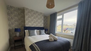 Een keurig opgemaakt bed met een blauw hoofdeinde, een bedlampje en een raam met gordijnen.
