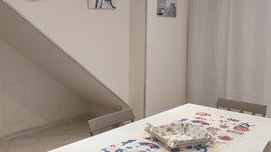 A dining area with a white table, chairs, and a table runner with nautical designs. There is a clock on the wall and a framed picture above the table. - At Erica's house (Mascali)