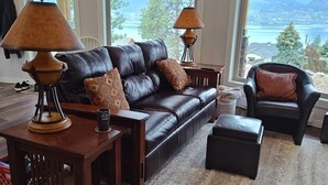 A living room with a leather sofa, two armchairs, a coffee table, and lamps. Large windows offer a view of a lake and mountains. - Private Suite with Amazing Lake View (Naramata)