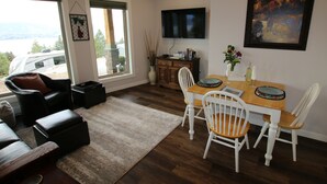 A living room with a sofa, a dining table with chairs, a TV mounted on the wall, and a window with a view of a vehicle and landscape. - Private Suite with Amazing Lake View (Naramata)