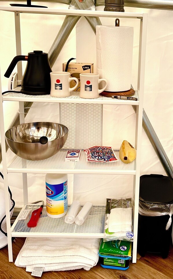 Un estante metálico blanco con varios objetos, incluyendo una tetera, una caja de filtros de café, tazas, un tazón, barajas de cartas, un envase de toallitas húmedas, un encendedor rojo y un cubo de basura.
