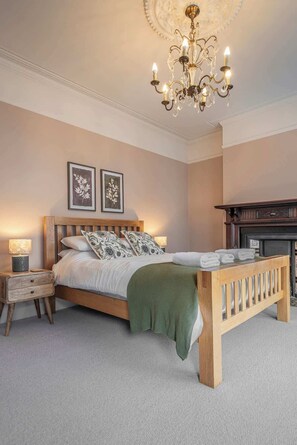 A bedroom with a wooden bed, a green throw blanket, a wooden bench, a fireplace, and two wall-mounted lamps.