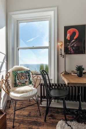 Ein Zimmer mit großem Fenster, einem Sessel mit Kissen mit Tiger-Muster, einem Tisch, einem Heizkörper und einer wandmontierten Leuchte.