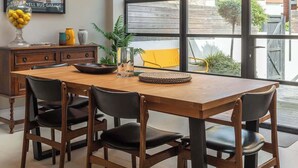 Un espace de salle à manger moderne, avec une table et des chaises en bois, un luminaire suspendu et une vue dégagée grâce à de grandes fenêtres.