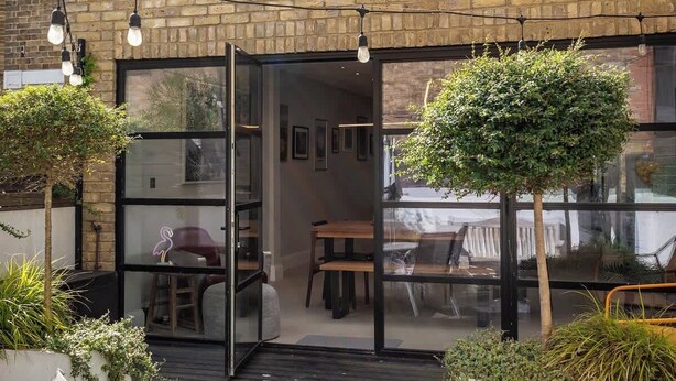 Un bâtiment en briques avec une entrée dotée d’une porte en verre, des places assises à l’extérieur et un petit arbre.