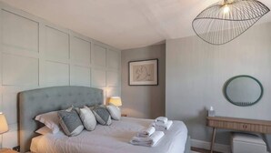 Un lit bien fait, agrémenté d’oreillers et de serviettes, une lampe de chevet, une œuvre d’art encadrée au mur et un miroir rond posé sur une console en bois.