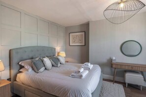 A neatly made bed with pillows and towels, a bedside lamp, a framed artwork on the wall, and a round mirror on a wooden console.