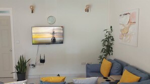 A living room with a TV mounted on the wall, a potted plant, a sofa with yellow cushions, and a painting on the wall. - Newly constructed luxury apartment. -Pristine Stay- (Roseau)