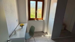 Ein kleiner, heller Raum mit Holzboden, einem Fenster mit Blick ins Grüne, einem Schreibtisch mit Stuhl und einem Bett mit gestreifter Decke.