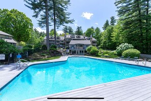 A swimming pool with a wooden deck, surrounded by trees and a multi-story building with balconies. - NEW! Charming Townhouse + Shared POOL (Gilford)