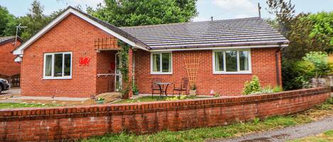 A single-story brick house with a sloped roof, a front porch, and a small garden area with potted plants and a wooden table.