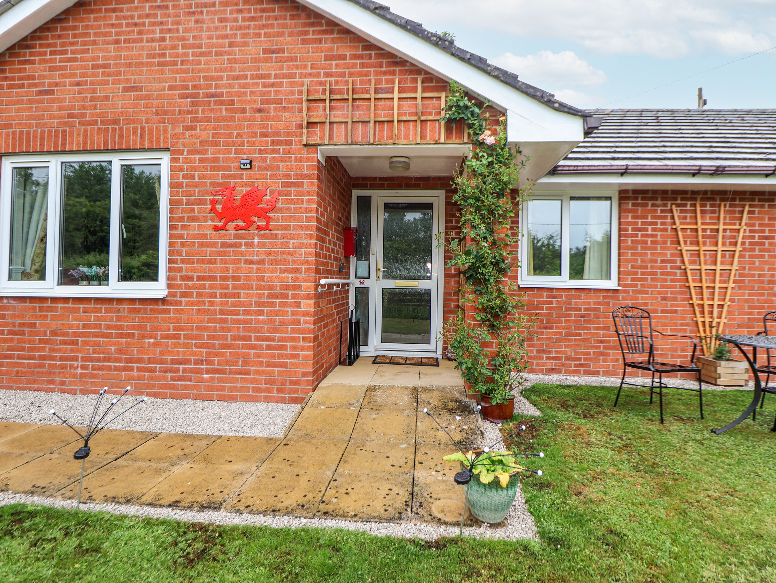 Une maison en briques rouges avec une porte blanche, un emblème représentant un dragon rouge et un petit jardin avec une table et des chaises.