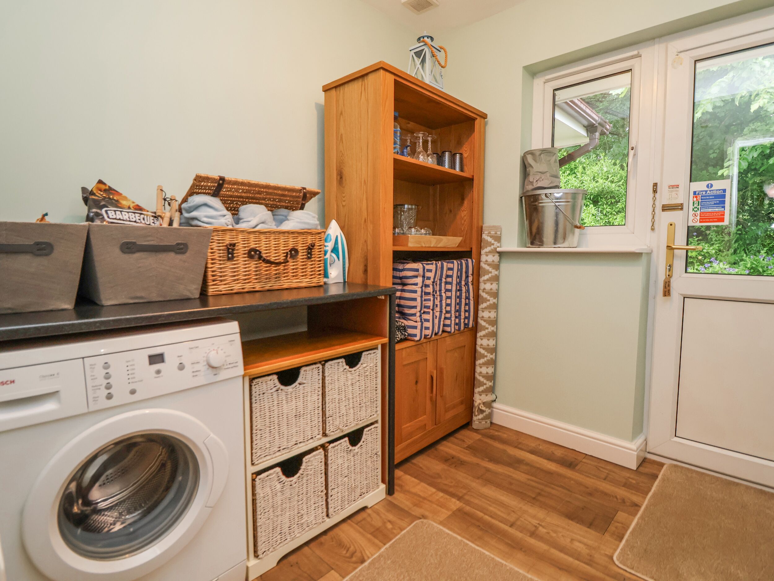 Un espace buanderie comprenant un lave-linge, des paniers en osier, une étagère en bois avec des produits de nettoyage et une fenêtre donnant sur un paysage verdoyant.