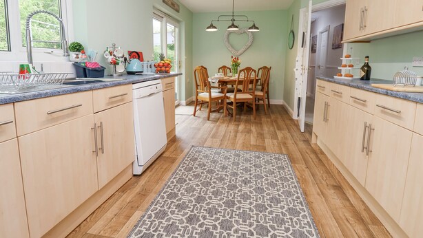 A kitchen with wooden cabinets, a dishwasher, a dining table with chairs, and a patterned rug. - 2 Mountain View (Wrexham)