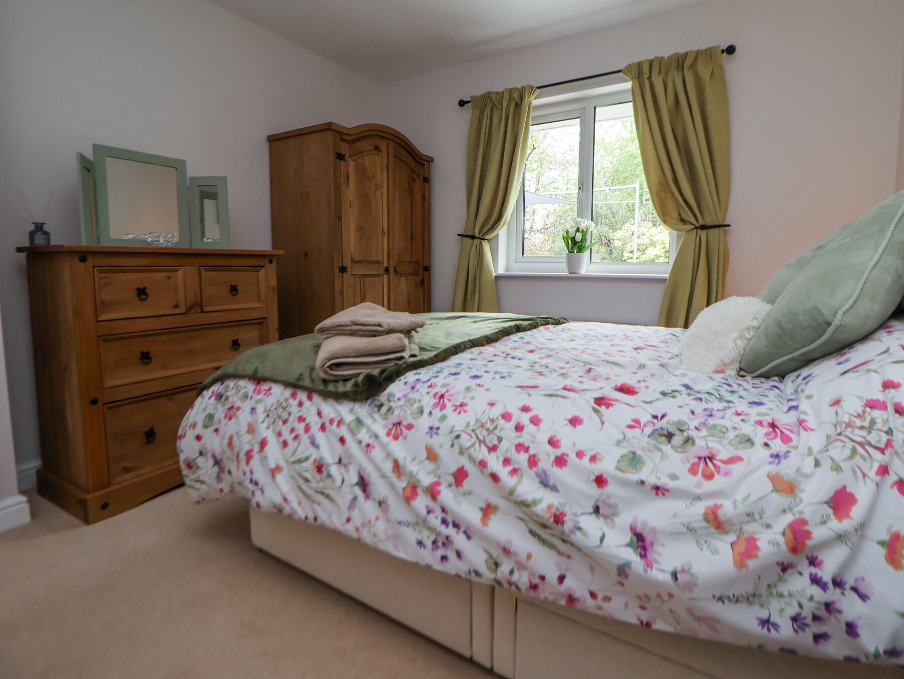 A bedroom with a floral bedspread, a wooden dresser, and a window with curtains.