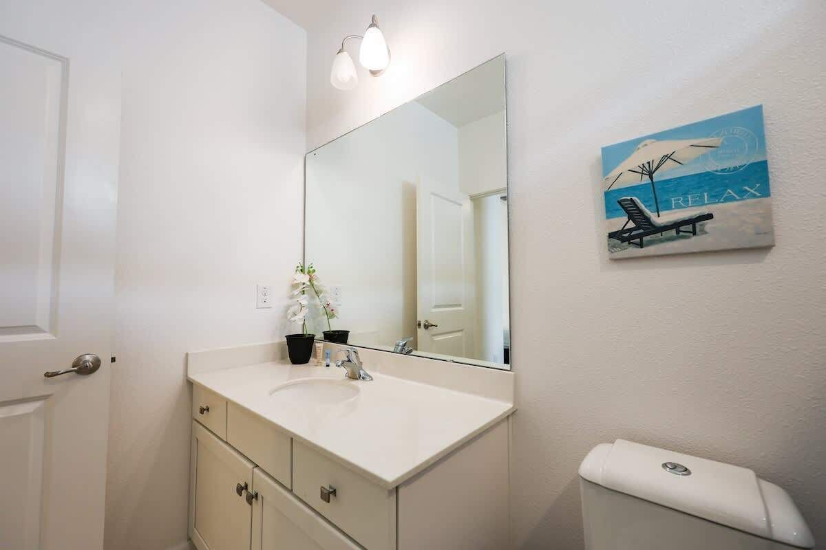 Une salle de bain avec un lavabo blanc, un grand miroir, des toilettes et une œuvre d’art fixée au mur représentant une scène de plage.