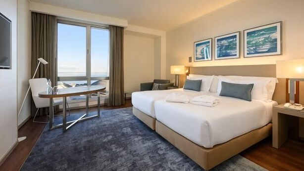 Een hotelkamer met een bed, een bureau, een stoel, een nachtkastje en uitzicht op zee.