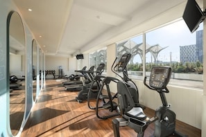 Une salle de sport équipée de tapis de course et d’appareils d’exercice, avec de grandes fenêtres et une vue sur un immeuble.