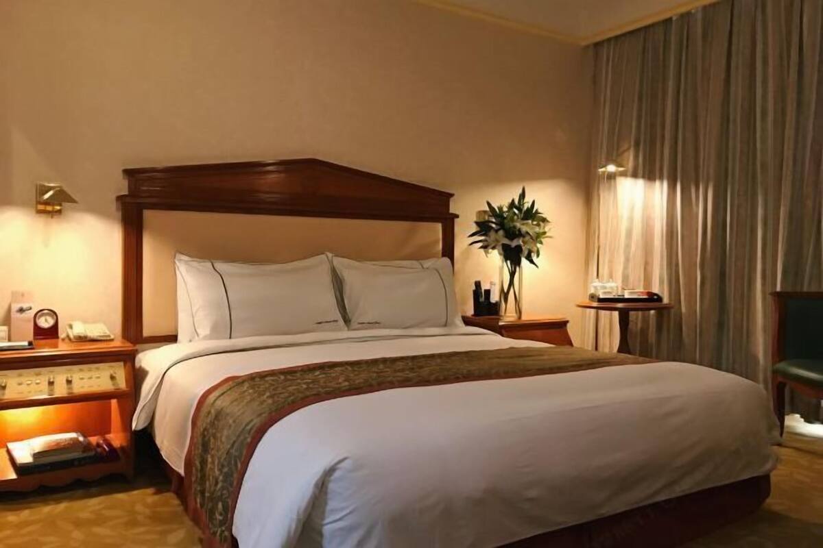 A hotel room with a large bed, a bedside table with a lamp, a clock, and a vase with flowers.
