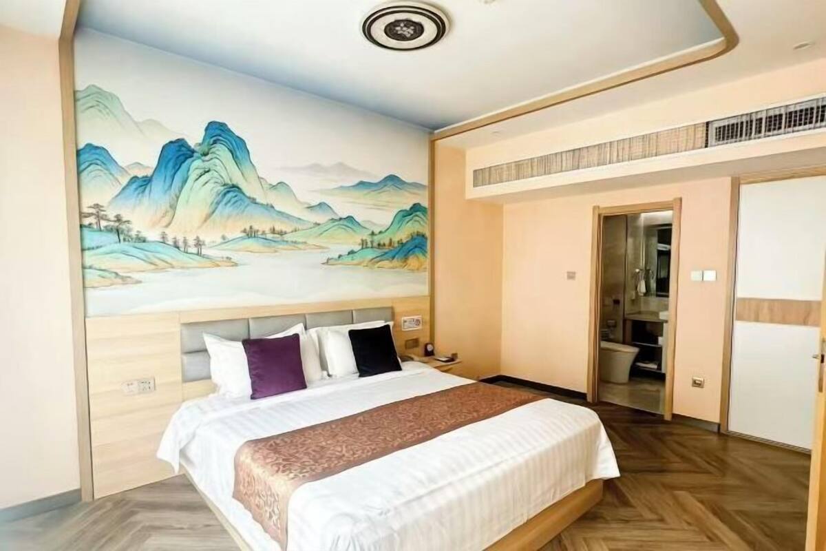 A hotel room with a bed, a painting of mountains on the wall, a bathroom visible through an open door, and a ceiling vent.