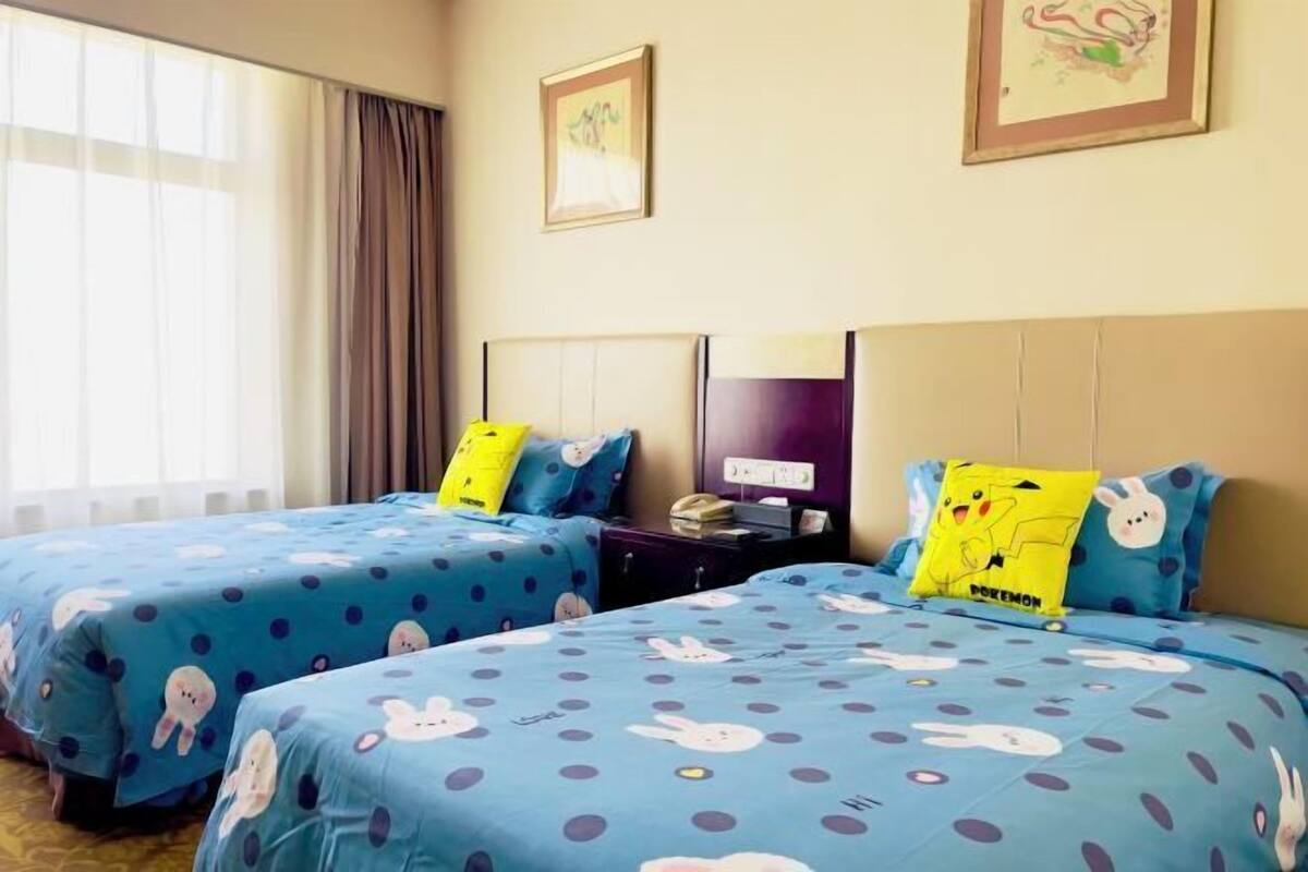 A hotel room with two beds, each with blue bedding featuring cartoon characters, a nightstand with a lamp, and a window with curtains.