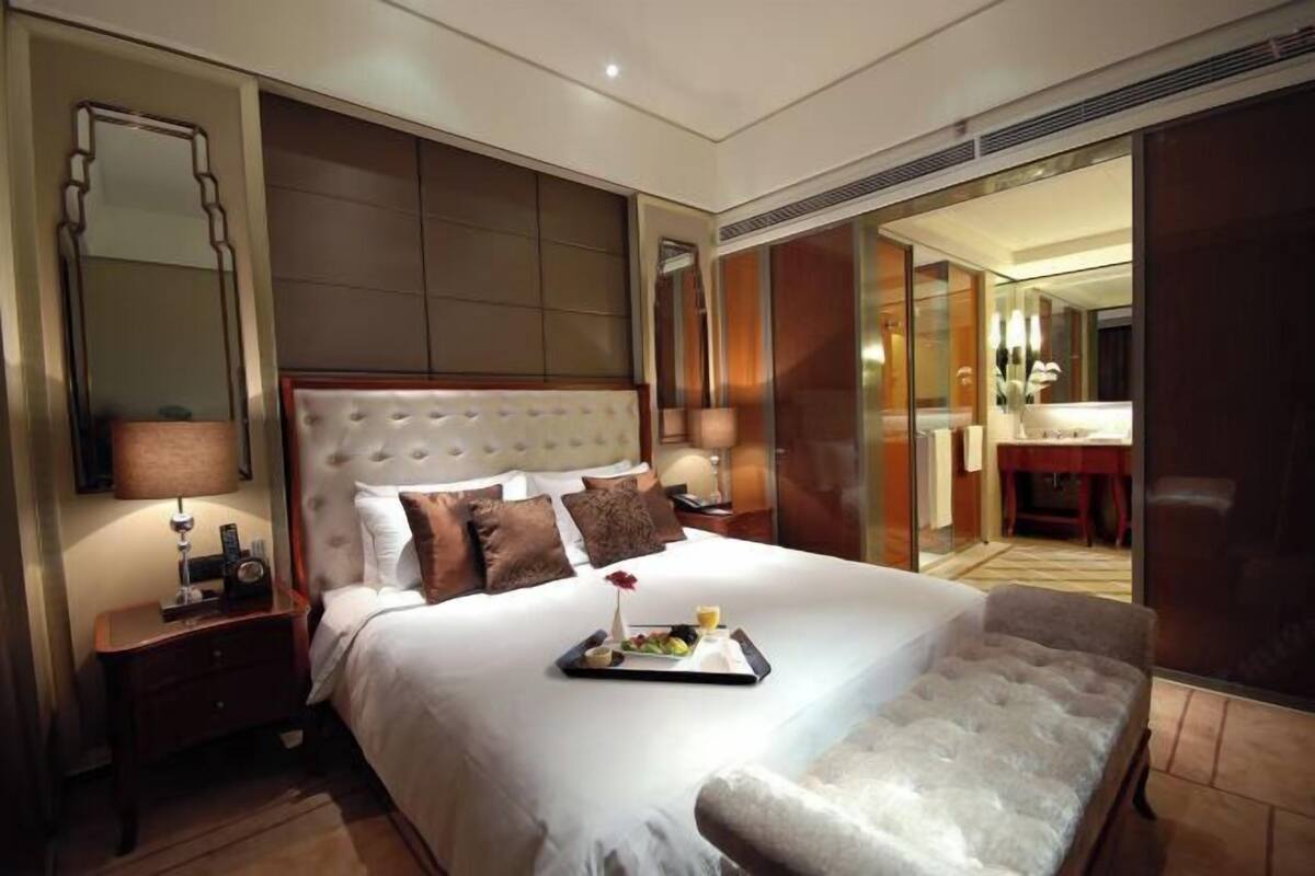 A hotel room with a large bed, a bedside table with a lamp, a tray with fruits and a drink, and a mirror on the wall.